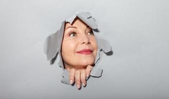 Surprised woman looking playfully in torn paper hole, has excited cheerful expression, looks through breakthrough of gray background. Wow, great news photo