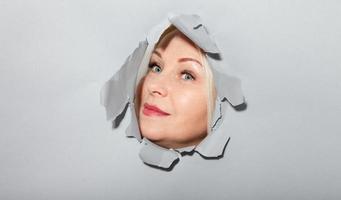 Surprised woman looking playfully in torn paper hole, has excited cheerful expression, looks through breakthrough of gray background. Wow, great news photo