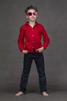 vertical longitud total retrato de un elegante pequeño chico en rojo Gafas de sol en un gris antecedentes. foto