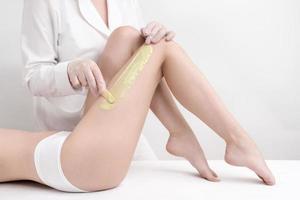 Cosmetologist applying green hot wax on slim woman leg using spatula while woman lying down on couch photo