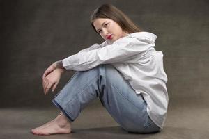 un hermosa niña en un blanco camisa y pantalones se sienta en un gris antecedentes. foto