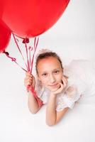 Girl holding red balloons in hand and positive looking at camera, lying down on white background