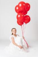 Smiling girl in white dress holding lot of red balloons in hand looking at camera with happy emotion