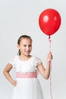 niña en blanco vestir participación rojo aire globos en mano, contento sonriente mirando a cámara. frente ver foto