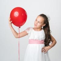 Portrait of girl in white dress holding one red balloon in hand and looking at balloon. Studio shot photo