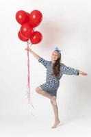 Ecstatic active girl holding bunch of red balloons in hand, standing on one leg on white background photo