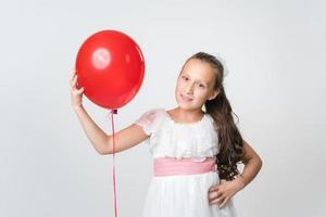 Portrait of happy girl in white dress holding one red balloon in hand, smiling and looking at camera photo