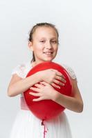 retrato sonriente niña abrazando rojo globo con ambos manos, mirando a cámara en blanco antecedentes foto