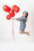 niña en saltar participación grande manojo de rojo globos en uno mano y sonrisas felizmente mirando a cámara foto