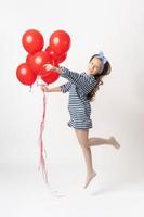 Playful girl in striped dress is jumping, holding bunch of red balloons in hand, looking at camera photo