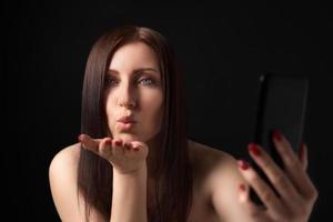 Woman blowing lips sending air kisses over palm, taking selfie to mobile phone on black background photo