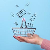 A woman's hand holds an empty shopping cart and icons of cards, coupons on a blue background. Sale and discount concept photo