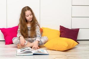 niña con fluido pelo en pijama en el piso entre multicolor almohadas con entusiasmo lee un libro foto