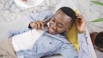 contento y linda africano joven hombre acostado en sofá a hogar y hablando en el teléfono. elegante y carismático africano joven hombre hablando en el teléfono a hogar. video