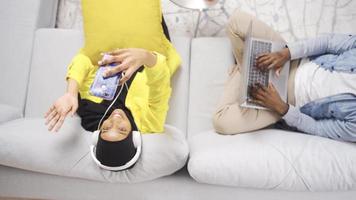Cheerful and happy African married couple sitting together on sofa and busy with technology gadgets. African man and hijab african woman listening to music with headphones at home using laptop. video