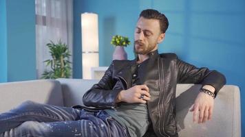 Cool and stylish adult young man sitting on sofa at home and getting lost in his deep thoughts. Stylish young man resting on sofa at home and thinking about plans, ideas, problems in mind. video