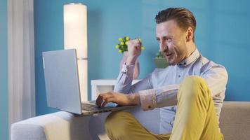 à moda e carismático adulto jovem homem usando computador portátil às casa alegria, obtendo Boa notícias, ganhando. elegante bonito homem sentado em sofá às casa e usando computador portátil alegria às o que ele vê em tela. video
