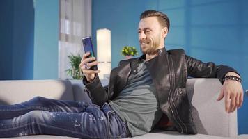 contento e bello giovane uomo video chat in linea, sorridente, godendo utilizzando il suo smartphone a casa. carismatico e elegante giovane uomo rilassante su divano a casa e video chat su Telefono.