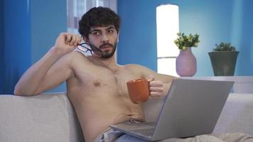Handsome young man with curly hair using laptop is thoughtful and naked. Attractive Young Man Thinking, Working From Home, Shopping Online, Watching Videos or Writing Emails.