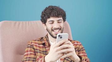 feliz jovem bonito homem sentado em sofá usando conectados Móvel aplicativos. sorridente jovem homem goza comunicando com amigos em social meios de comunicação ou jogando jogos em a telefone. video