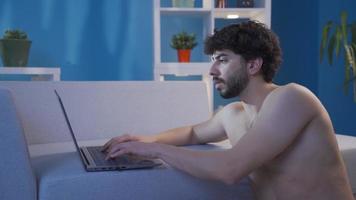 nudo bello giovane uomo utilizzando il computer portatile a casa a notte. giovane nudo bello uomo seduta su il pavimento a casa utilizzando il computer portatile. video