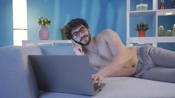 Young man using laptop at home at night talking on the phone. Nude male model. Young naked man lying on sofa alone at home using laptop and talking on the phone. video