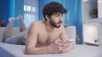 Young male model lying naked on sofa and texting using his smartphone. Young man with attractive and fit body texting with girls on the phone. video