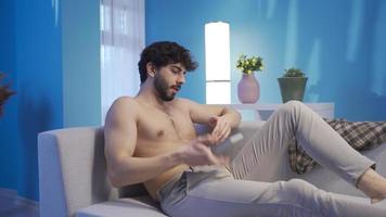 Naked young man texting on the phone and getting angry. Young man with a healthy fit body is lying on the sofa texting on the phone and receiving news that will upset him. video