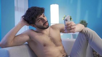 desnudo y atractivo joven hombre utilizando su teléfono inteligente hermoso joven hombre con un ajuste y ajuste cuerpo acostado felizmente en el sofá, mirando a su teléfono. video