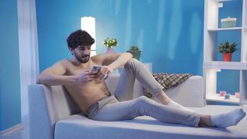 Handsome young man with a fit and fit body is lying happily on the sofa, looking at his phone. Male model with handsome and fit body looking at his smartphone lying on the sofa.