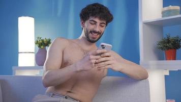 Jeune homme avec nu génial physique à la recherche à le sien intelligent téléphone seul à maison, SMS, en utilisant social médias. video