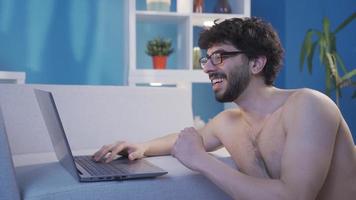 Young naked man using laptop rejoices, wins, triumphs, laughs. Naked young man in glasses using laptop alone at home, typing on keyboard rejoicing and being happy. video