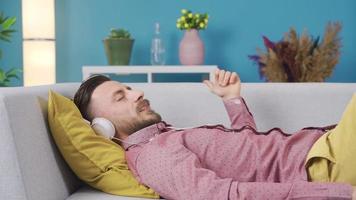 alegre y elegante hombre escuchando a música y descansando a hogar. hombre relajante en sofá a hogar con auriculares escuchando a música, soñador y contento. video