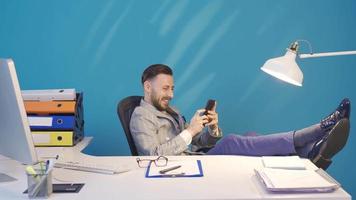 Relaxed businessman stretches his feet on the table and uses the phone. Happy and carefree businessman enjoying his desk with his feet up and using his smart phone. video