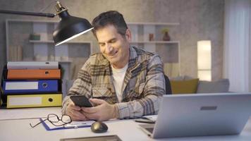 souriant mature homme travail dans Accueil Bureau à la recherche à téléphone, SMS, surfant social médias et étant heureux. mature homme en utilisant intelligent mobile téléphone souriant, prise une Pause de travail. video