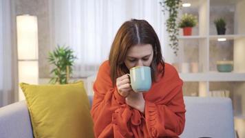 enfermo y agotado joven mujer sentado en sofá en vivo habitación a hogar y Bebiendo caliente café o sano bebidas a calentar arriba. video