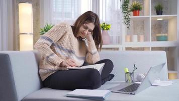 Young college girl resting and studying on sofa at home. Calm and relaxed young woman sitting on sofa at home and working on laptop and studying her studies taking notes on paper video