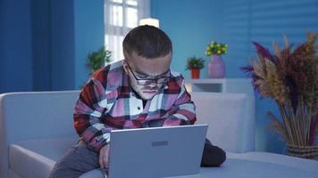 Young man with dwarfism doing focused and serious work at home at night using laptop. Disabled dwarf young man sitting on sofa alone at home using laptop, doing serious. video