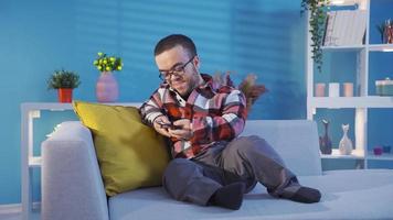 Inaktiverad dvärg- ung man vilar på soffa på Hem och ser på hans smartphone. Inaktiverad dvärg- ung man Sammanträde på soffa på Hem vilar och använder sig av telefon. video
