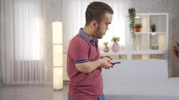 Happy dwarf man looking at his phone at home, texting, reading news, shopping online. Young dwarf man sitting on sofa at home and using smartphone. video