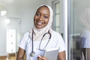 Young female musllim African clinician doctor in scrubs using touchpad while communicating with patients online photo