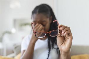 African girl in glasses rubs her eyes, suffering from tired eyes, ocular diseases concept. young woman holds glasses with diopter lenses and rubs her eyes from fatigue fatigue of vision photo