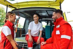 paramédicos y médico en pie en el lado ambulancia. médico es que lleva un médico trauma bolsa. grupo de Tres paramédicos en pie en frente de ambulancia con sonrisa. foto