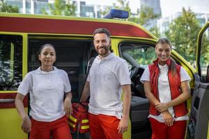 Three multiracial paramedics standing in front of ambulance vehicle, carrying portable equipment photo