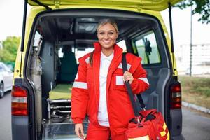 Young woman , a paramedic, standing at the rear of an ambulance, by the open doors. She is looking at the camera with a confident expression, smiling, carrying a medical trauma bag on her shoulder. photo