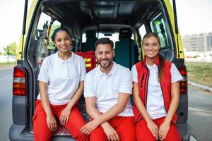 paramédicos y médico en pie a posterior de ambulancia. médico es que lleva un médico trauma bolsa. grupo de Tres paramédicos en pie en frente de ambulancia con sonrisa. foto