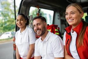 paramédicos y médico sentado a posterior de ambulancia. grupo de Tres paramédicos en pie en frente de ambulancia con sonrisa. foto