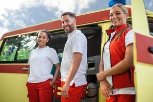 Three multiracial paramedics standing in front of ambulance vehicle, carrying portable equipment photo