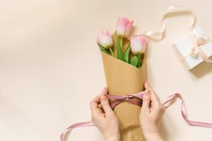 De las mujeres manos paquete un ramo de flores de Fresco tulipanes utilizando marrón papel ecológico y satín cinta, flatlay composición en un beige antecedentes. foto