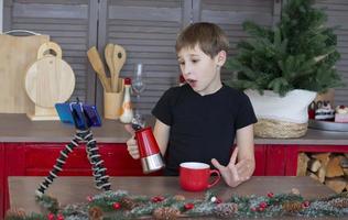 A portrait of a confident and creative child blogger talking to the camera at home in the kitchen, he is vlogging. A little boy is recording a stream on a smartphone. photo